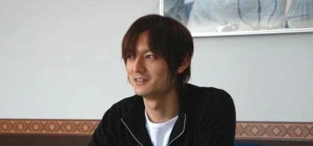 Jun Maeda — นักเขียนบท นักแต่งเพลง และผู้ร่วมก่อตั้ง Key Studios Jun Maeda ในงานสาธารณะ นั่งอยู่หน้าไมโครโฟนหันหน้าเข้าหาผู้ชม