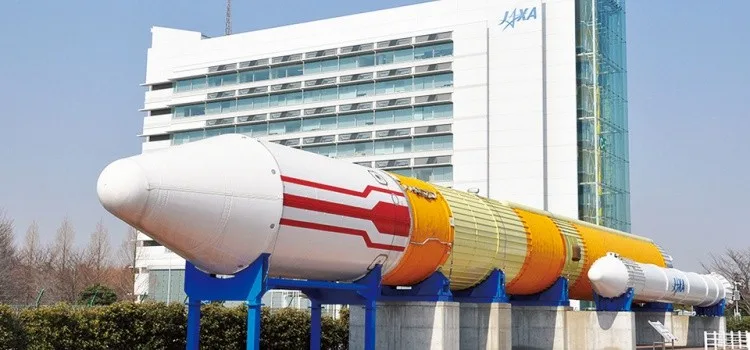 Logo e lettering della JAXA sulla parete di un edificio, con un razzo sulla rampa di lancio sullo sfondo