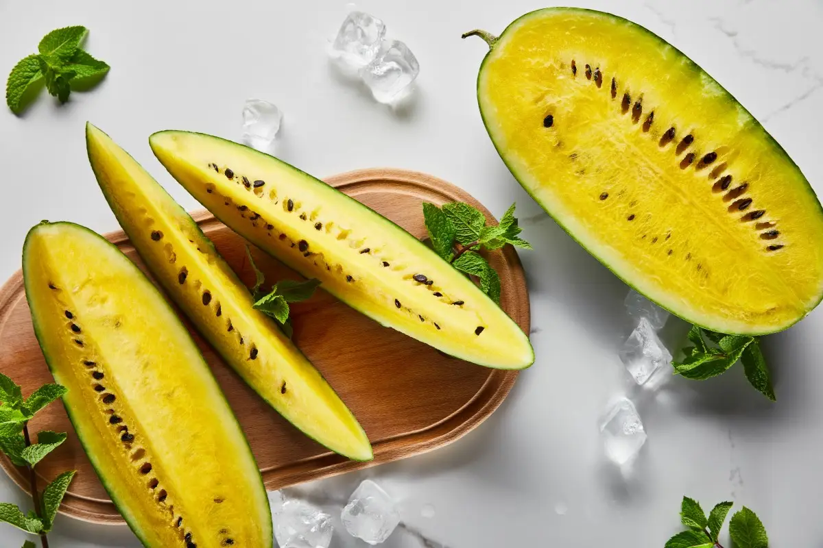 top view of cut delicious exotic yellow watermelon with seeds on marble surface with mint, ice and wooden chopping board