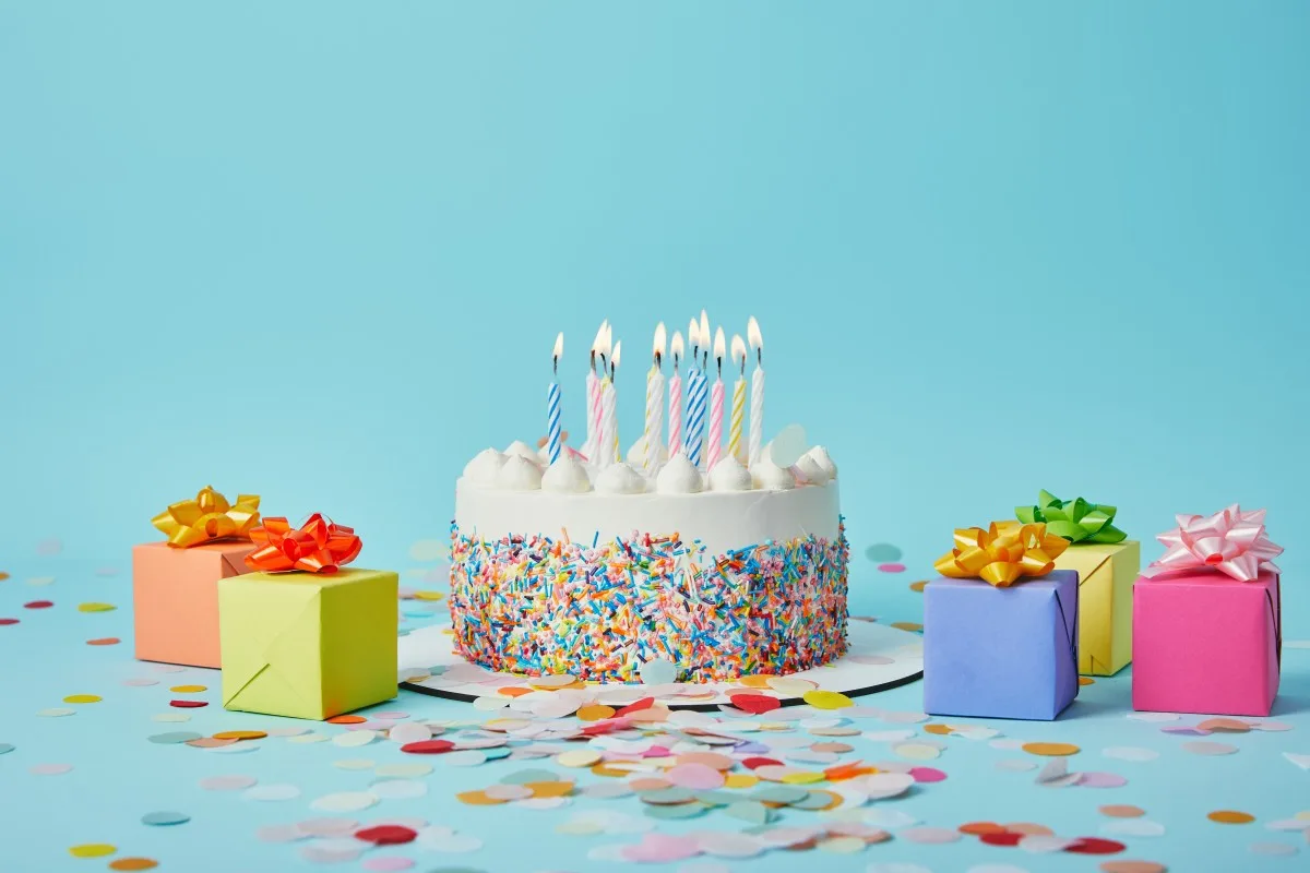 Delicious cake with candles, colorful gifts and confetti on blue background