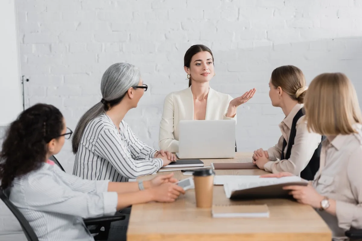 happy team leader pointing with hand near laptop and multicultural businesswomen during meeting